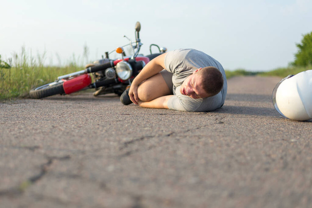 Een man houdt vast aan zijn knie, liggend op het asfalt in de buurt van zijn motor, het thema van verkeersongevallen - Foto, afbeelding