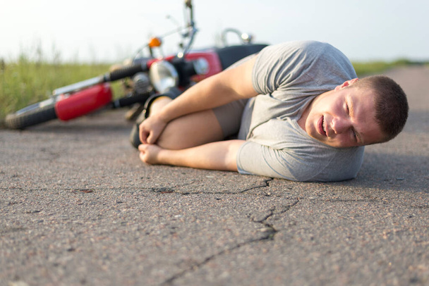 Ένας άνθρωπος κρατά το γόνατό του, που βρίσκεται στην άσφαλτο κοντά στη μοτοσικλέτα του, το θέμα των τροχαίων ατυχημάτων - Φωτογραφία, εικόνα