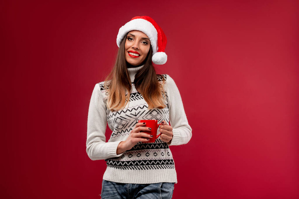 Close up portret prachtige blanke vrouw in rood Santa hoed op rode studio achtergrond. Kerstmis Nieuwjaar vakantie concept. Schattig meisje tanden glimlachen positieve emoties met rode kop koffie in handen - Foto, afbeelding
