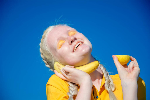 A young albino woman holds a banana to her ear like a phone. against the blue sky. - Fotó, kép