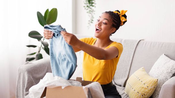 Счастливая чернокожая женщина распаковывает одежду после интернет-покупок - Фото, изображение