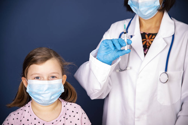 Children vaccinating immunization concept. Doctor injecting vaccination in arm of a little girl with face mask. - Фото, изображение