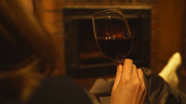Woman with glass of red wine is resting by the fireplace. - Foto, afbeelding