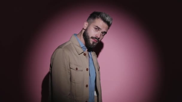 dramatische jonge kerel naar beneden kijken, poseren in een zijaanzicht positie, vaststelling beige jas en spinnen in een mode licht op roze achtergrond in studio - Video