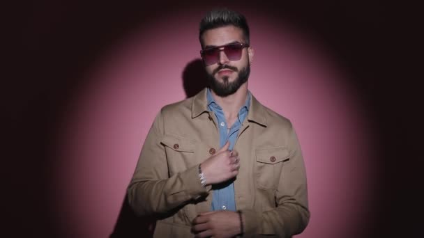 cool young man in blue shirt wearing sunglasses and arranging beige jacket, taking off glasses and sensually making silence gesture in a fashion light on pink background in studio - Footage, Video