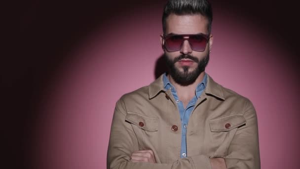 serieuze jongeman in denim shirt dragen zonnebril en beige jas kruising armen en knikken in een mode licht op roze achtergrond in studio - Video