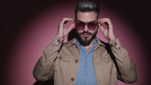 fashion cool guy in denim shirt adjusting beige jacket, taking off glasses and posing, confidently crossing arms in a fashion light on pink background in studio - Footage, Video