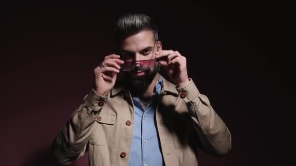 cool fashion model in denim shirt zetten zonnebril op, het regelen van beige jas en vol vertrouwen kruisende armen in een schaduw op roze achtergrond in studio - Video