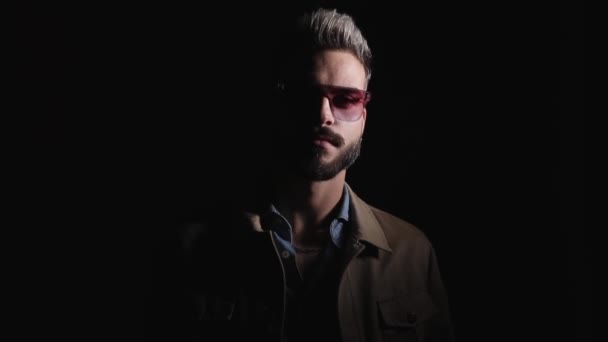 cool unshaved man coming from the dark, sensually fixing collar and jacket, looking to side and arranging glasses, confidently making gun gesture and leaving on black background in studio - Footage, Video