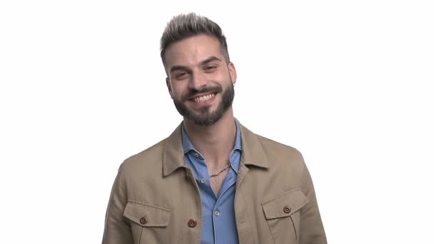 felice giovane uomo in camicia di jeans divertirsi e ridere, regolazione giacca beige e braccia incrociate, in piedi isolato su sfondo bianco in studio - Filmati, video