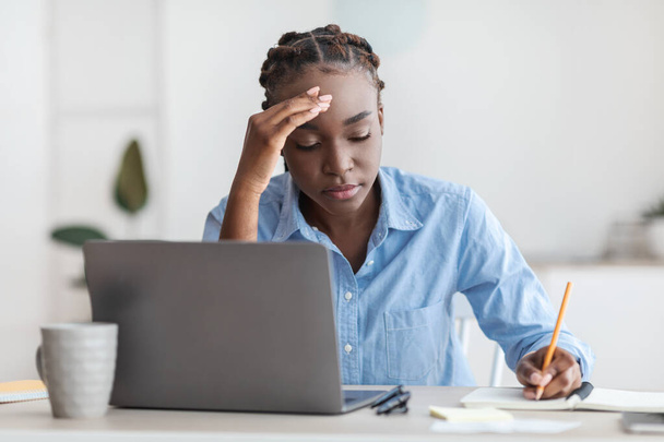 Tired black female office employee sitting at desk with laptop, taking notes - Zdjęcie, obraz