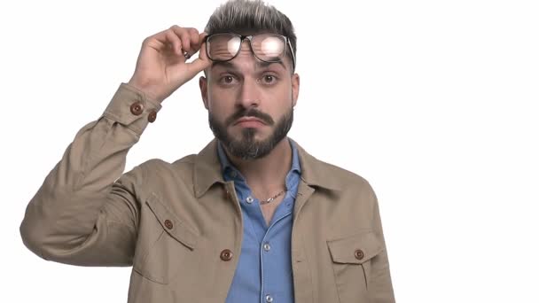 young casual man in beige jacket putting and taking off glasses, changing emotions, sticking out tongue, smiling, laughing, making a crazy hand gesture, pointing fingers to temple and standing isolated on white background - Footage, Video