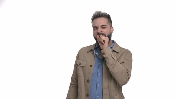 sonnolento giovane uomo che indossa giacca beige tenendo pugno a mano e sbadigliando, in piedi isolato su sfondo bianco in studio - Filmati, video