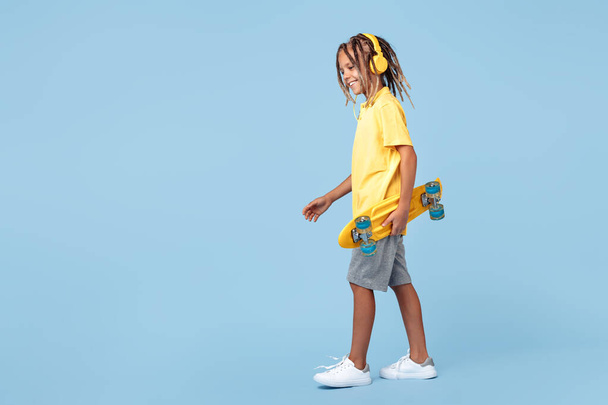 Modern little boy with african dreads with headphone and skateboard posing over blue background. - Foto, Imagen