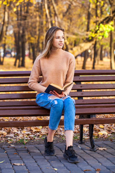 Beautiful young girl with a book sits on a bench in an autumn park - Foto, Imagem