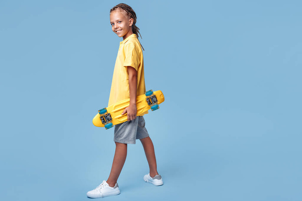 Full lengh image of stylish little boy with african dreads with headphone and skateboard posing over blue background. - Valokuva, kuva