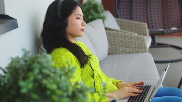 Fiatal ázsiai nő, szabadúszó, fejhallgatóval dolgozik a laptopján. Távolsági tanulás vagy távmunka fogalma - Felvétel, videó