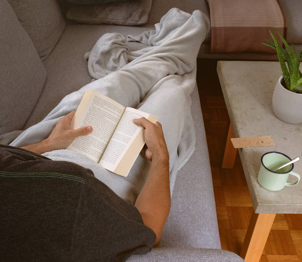 Mann in gemütlicher Wohnung, eingewickelt in eine Decke, entspannt sich auf Sofa, Buch in der Hand und liest. Warmes Sonnenlicht ins Wohnzimmer - Foto, Bild