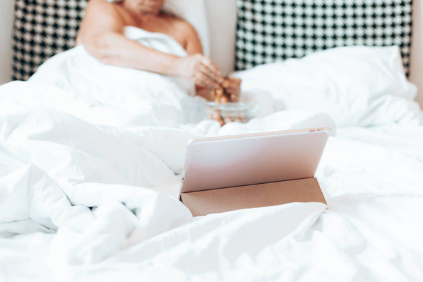 自宅でベッドの中でタブレットコンピュータを持つブロンドの女性は、映画やインターネットを見て。ブドウの実を食べなさい。おはようございます - 写真・画像