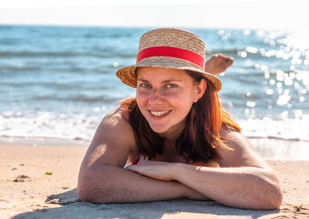 Young girl in a hat and swimsuit lies on the beach, looking at the camera - Photo, Image