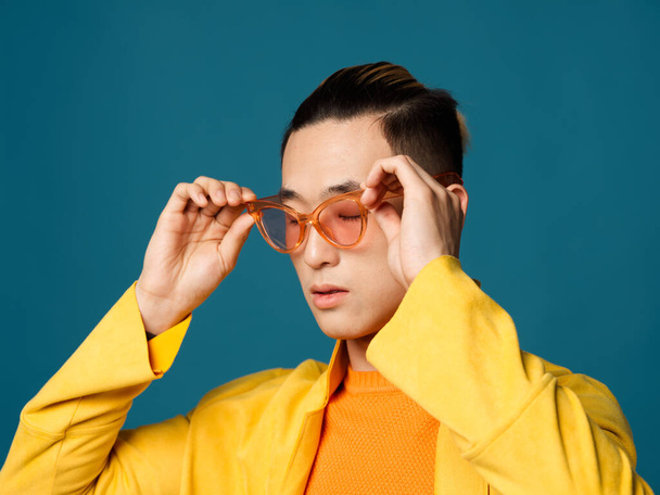 Asian man straightens glasses on his face and yellow jacket blue background - Photo, Image