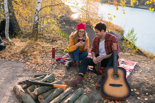 秋の木々や川を背景に、カップルが自然の中で休んでいます。男はギターを弾く - 写真・画像