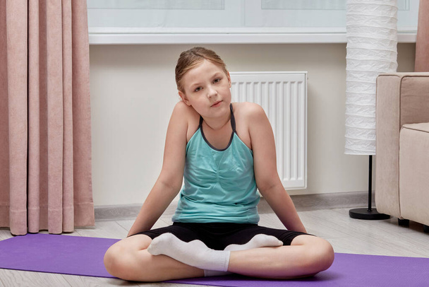 Beautiful girl sitting on a purple sports Mat doing yoga. Looks at the camera. - Foto, afbeelding