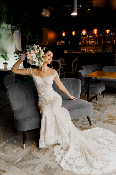 Bride in wedding dress and bridal veil seats on fashion chair in cafe. Advert for social networks for wedding agency and bridal salon - Foto, imagen