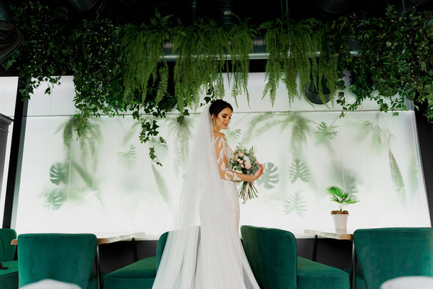 Young attractive bride in wedding dress with bridal veil looks at bouquet and smiles. Girl in wedding gown in modern restaurant. Advert for social networks for wedding agency. - Fotografie, Obrázek