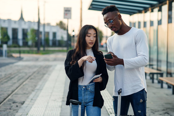 Handsome african american man and asian pretty girl go to business trip with suitcases and looking map at smartphone. - Foto, immagini