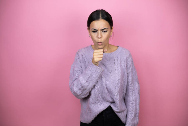 Schöne Frau trägt einen lässigen violetten Pullover über rosa Hintergrund mit der Hand vor dem Mund, weil sie hustet - Foto, Bild
