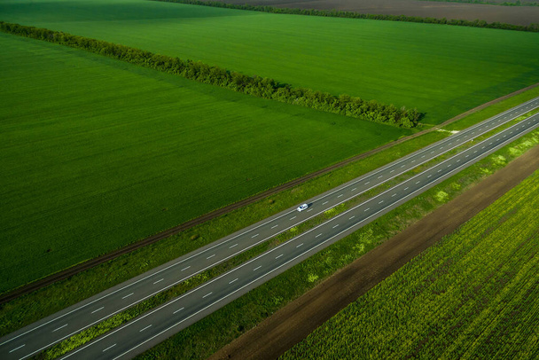 白い車が緑の畑に沿ってアスファルトの道路を走行しています。空気中から見た。空中風景。ドローン撮影。旅行のコンセプト - 写真・画像