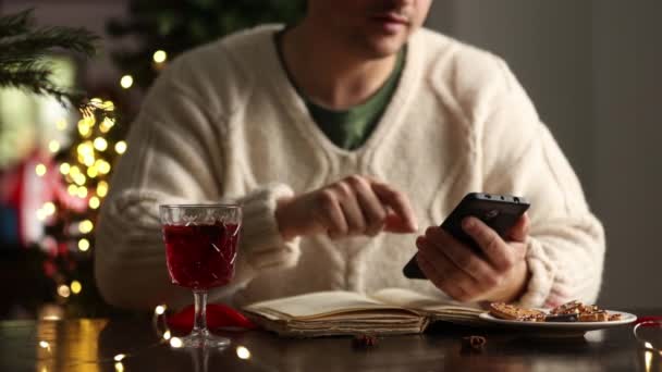 Ο άνθρωπος κάθεται στο τραπέζι με το κινητό τηλέφωνο και πίνοντας ένα ζεστό κρασί  - Πλάνα, βίντεο