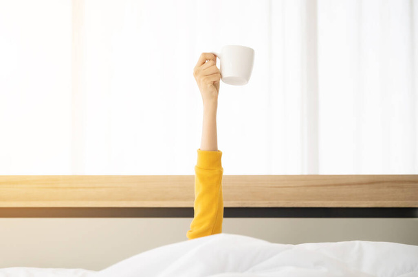 Woman waking up in the morning on bed. She hiding under the blanket and stretching out arms with holding a cup of coffee - Photo, Image