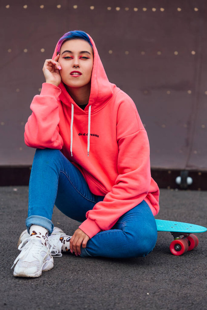 Portrait of a trendy pretty young girl dressed in pink hoodie and jeans sitting next to the skateboard court on her blue plastic skateboard. - Foto, afbeelding