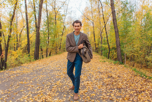 Tall handsome man dressed in a brown jacket walking with a bag on the autumn alley - Foto, Imagen