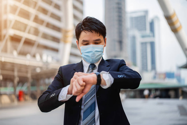 business man checking time on his watch and wearing medical mask during coronavirus (covid-19) pandemic in the city - 写真・画像