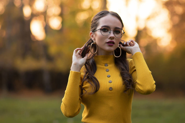 Junge langhaarige Brünette in gelbem Strickpullover und Brille blickt in die Kamera, während sie im Park vor dem verschwommenen Laub der Herbstbäume steht - Foto, Bild