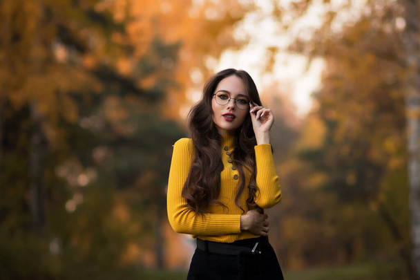 Junge langhaarige Brünette in gelbem Strickpullover und Brille blickt in die Kamera, während sie im Park vor dem verschwommenen Laub der Herbstbäume steht - Foto, Bild