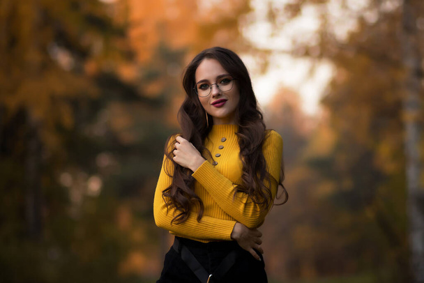 Młoda brunetka z długimi włosami w żółtych dzianinach sweter i okulary patrząc w aparat, stojąc przed rozmazanymi liśćmi jesiennych drzew w parku - Zdjęcie, obraz