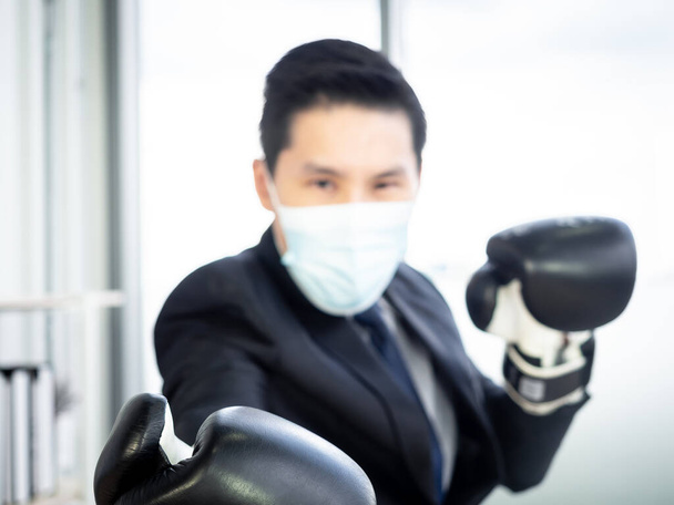 Blurred of Asian Businessman in suit and protective face mask and focus on hand wearing boxing gloves in office. Man with challenge wears mitten, posture in boxing ready to fight the problem concept. - Photo, Image