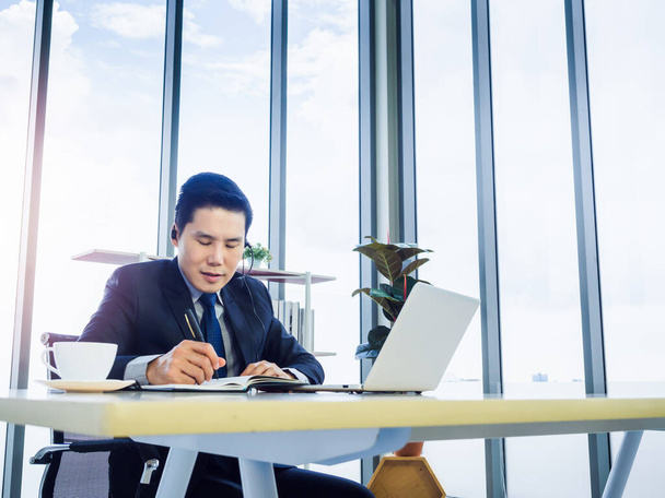 Ázsiai üzletember öltönyt visel fejhallgatóval jegyzetel, és laptopot használ az irodában, közel a hatalmas üvegablakhoz és az égbolt hátteréhez. Konferenciahívás és ügyfélszolgálati üzemeltetői koncepció. - Fotó, kép