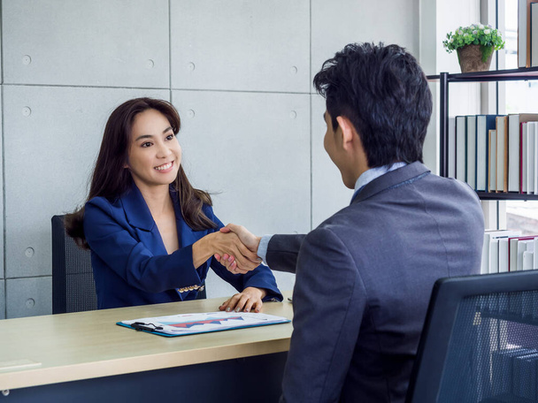 Aziatische zakenvrouw en zakenman in pak die elkaar de hand schudden. glimlachende vrouwelijke manager zitten aan bureau in kantoor handdruk met jonge mannelijke werknemer na het werken samen. - Foto, afbeelding