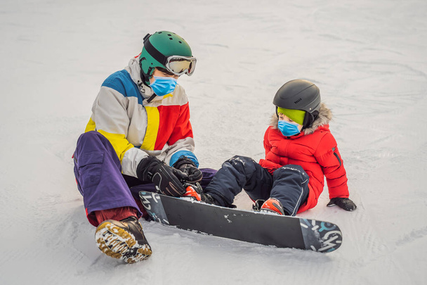 スノーボードインストラクターは、 COVID-19コロナウイルス中に医療マスクを着用してスノーボードに男の子を教えています。冬の子供たちのための活動。子供冬のスポーツ。ライフスタイル - 写真・画像