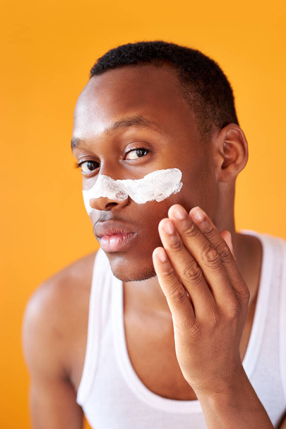 young african-american guy applying face cream under his eyes on yellow background - Φωτογραφία, εικόνα