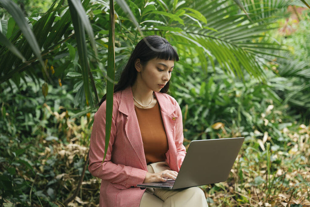 Mujer de negocios asiática en camisa formal sentada en una silla usando computadora portátil en la naturaleza. - Foto, imagen