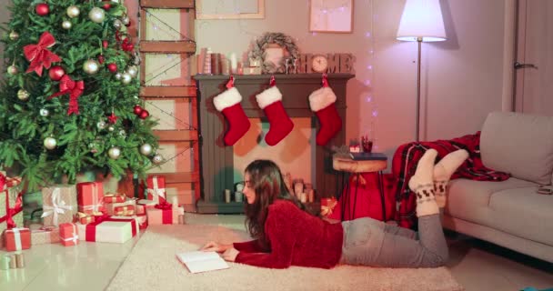 Женщина читает книгу у камина дома в канун Рождества - Кадры, видео