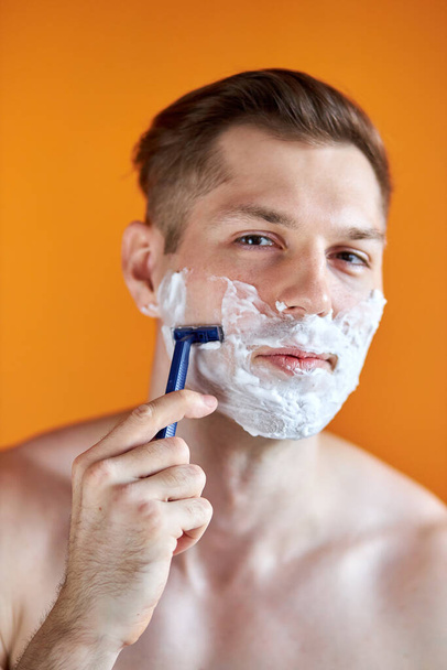 portrait of caucasian male shaving his face with foam and razor isolated on orange background - Fotografie, Obrázek
