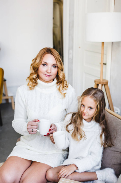 Mother and daughter in light sweaters are sitting on the sofa, the room is decorated for the new year and Christmas, hugs, conversations, smiles, kisses, waiting for a gift and magic - Фото, изображение