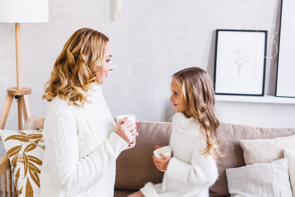 Mother and daughter in light sweaters are sitting on the sofa, the room is decorated for the new year and Christmas, hugs, conversations, smiles, kisses, waiting for a gift and magic - Foto, Imagem